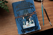 Blue notebook with 'Doctor Dolittle's Caravan' design on a wooden surface with writing utensils.