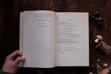 Open book on a wooden surface with a hand holding it, surrounded by small objects.