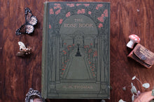 Decorative book titled 'The Rose Book' by A.H. Thomas on a wooden surface with decorative elements.