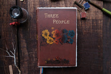 Vintage book titled 'Three People' with floral design on a wooden surface