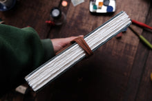 Hand holding a leather-bound notebook on a wooden surface with art supplies in the background