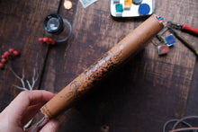 Decorative cylindrical object held by a hand on a wooden surface with art supplies.