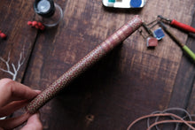 Hand holding a textured brown pen on a wooden surface with art supplies.