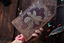 Hand holding a decorative leather-bound book with floral designs on a wooden surface.