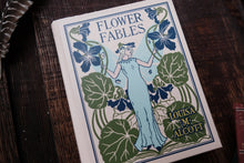 Book titled 'Flower Fables' by Louisa M. Alcott on a wooden surface with other books.