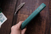 Hand holding a green book on a wooden surface with a feather and another book in the background.