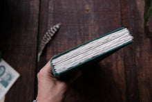 Hand holding a green-bound book on a wooden surface with a feather in the background