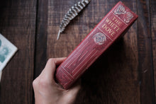 Red book titled 'The White Dove' held by a hand on a wooden surface with a feather and card.