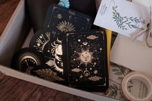 A styled flat-lay of a tarot deck inside a gray cardboard box, featuring black cards with intricate gold celestial illustrations. A white paper tag with green botanical illustrations and twine is tucked next to the deck, along with a roll of patterned tape on a wooden surface.