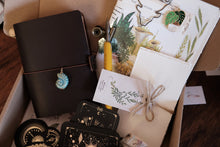 A curated gift box filled with bohemian-style items including a dark brown leather journal with a blue nautilus shell charm, botanical art prints, a deck of tarot cards, a small yellow-handled tool, and a twine-wrapped card on a wooden surface.