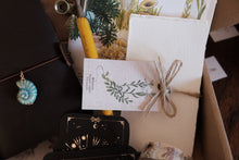 A styled flat-lay of a botanical-themed gift box or stationery set, featuring a white textured card with green leaf illustrations tied with twine, a yellow-handled tool, a black card holder with gold celestial designs, and a blue spiral shell charm.