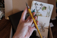 A hand holding a yellow-handled calligraphy brush over an open cardboard box containing art supplies, including printed cards and a black case.
