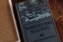 A vintage hardcover book titled 'Mother West Wind's Neighbors' by Thornton W. Burgess, featuring a dark gray cover with a black-and-white illustration of a frog in a pond, styled on a wooden surface next to another book.