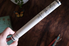 A hand holding a rolled-up cream-colored fabric scroll with black printed text, photographed over a dark wooden tabletop with blurred decorative objects in the background.