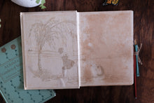 An open vintage-style sketchbook or journal on a dark wooden surface, showing faint sepia illustrations on the pages. A mint green book titled 'A Child's Garden of Verses' is partially visible underneath on the left, and two colored pencils are tied to the right edge with string.