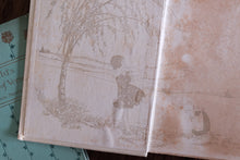 An open vintage book with aged, stained cream-colored pages featuring delicate sepia illustrations of a girl by a tree, resting on a teal book.