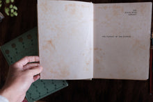 An open vintage book with aged, yellowed pages held by a hand, placed on a dark wooden surface next to another book.
