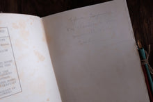 An open vintage book with handwritten inscription on the right page, photographed close-up on a dark wooden surface with a pencil tied to the edge.