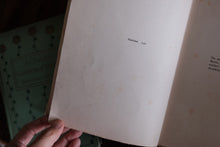A hand holding open a vintage book, showing blank aged cream pages with small printed text, with another book partially visible in the background.