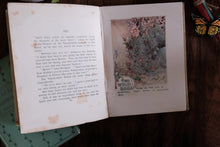 An open vintage book lying on a dark wooden surface, showing printed text on the left page and a floral illustration on the right page. A green notebook or book is partially visible underneath on the left, and a butterfly decoration appears in the top right corner.
