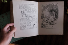 An open vintage book held by a hand, showing printed text and black-and-white illustrations on aged cream pages against a dark wooden background.