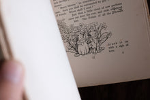 A close-up of an open vintage book showing printed text and a black-and-white illustration of a child and flower on the page, with a blurred hand holding the book.