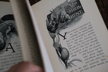 Open book with a black and white illustration held by a hand on a wooden surface.